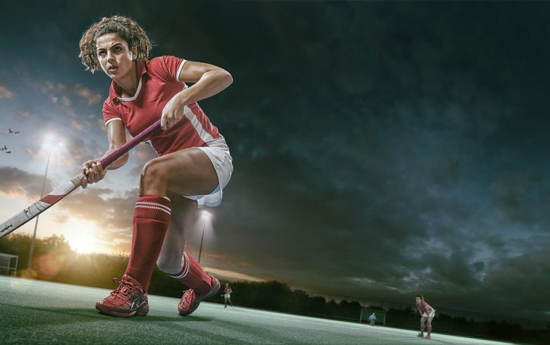A mid action low angle image of a female field hockey player dressed in generic red and white kit holding hockey stick about to strike ball during a game of field hockey. The action occurs in an floodlit artificial outdoor hockey uner a dramatic sky at sunset.
