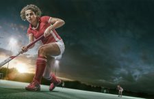 A mid action low angle image of a female field hockey player dressed in generic red and white kit holding hockey stick about to strike ball during a game of field hockey. The action occurs in an floodlit artificial outdoor hockey uner a dramatic sky at sunset.