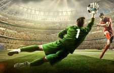 A male soccer player kicks a ball and soccer goalie jumping in motion for a ball while defending his gates on wide angle panoramic image of a outdoor soccer stadium or arena full of spectators under a sunny sky. The image has depth of field with the focus on the foreground part of the pitch. With intentional lensflares. Players are wearing unbranded soccer uniform.