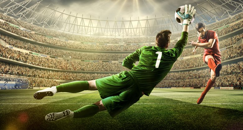 A male soccer player kicks a ball and soccer goalie jumping in motion for a ball while defending his gates on wide angle panoramic image of a outdoor soccer stadium or arena full of spectators under a sunny sky. The image has depth of field with the focus on the foreground part of the pitch. With intentional lensflares. Players are wearing unbranded soccer uniform.